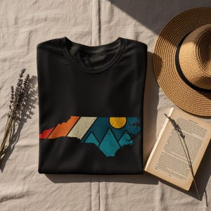 Black t-shirt with mountain and sun graphic, beside straw hat and book.
