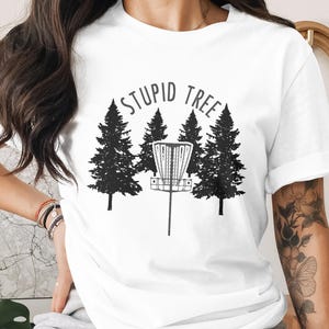 May include: White t-shirt with a black graphic of a disc golf basket and pine trees. The text "STUPID TREE" is arched above the basket. The shirt is made of a soft material and has rolled-up sleeves.