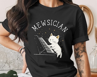 Könnte beinhalten: Schwarzes T-Shirt mit einer Illustration einer weißen Katze, die Klavier spielt, und dem Wort "MEWSICIAN" darüber. Das Design hat ein musikalisches Thema mit Sternen im Hintergrund. Ein skurriles und lustiges Design.