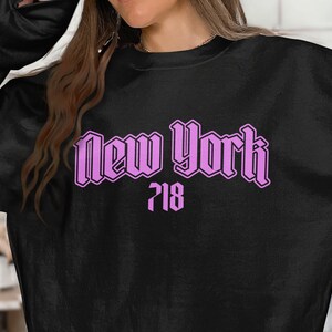 May include: Black sweatshirt with the words "New York" in a light purple, gothic-style font, with the number "718" below. The sweatshirt is a solid black color.