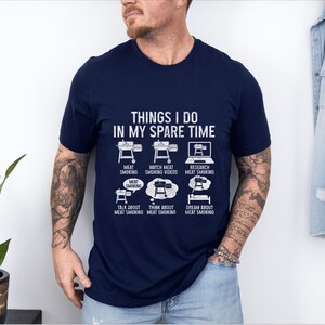 May include: Navy blue t-shirt with white text and graphics. The shirt reads "THINGS I DO IN MY SPARE TIME" with illustrations of a smoker, laptop, and thought bubbles. The graphics depict activities related to meat smoking.