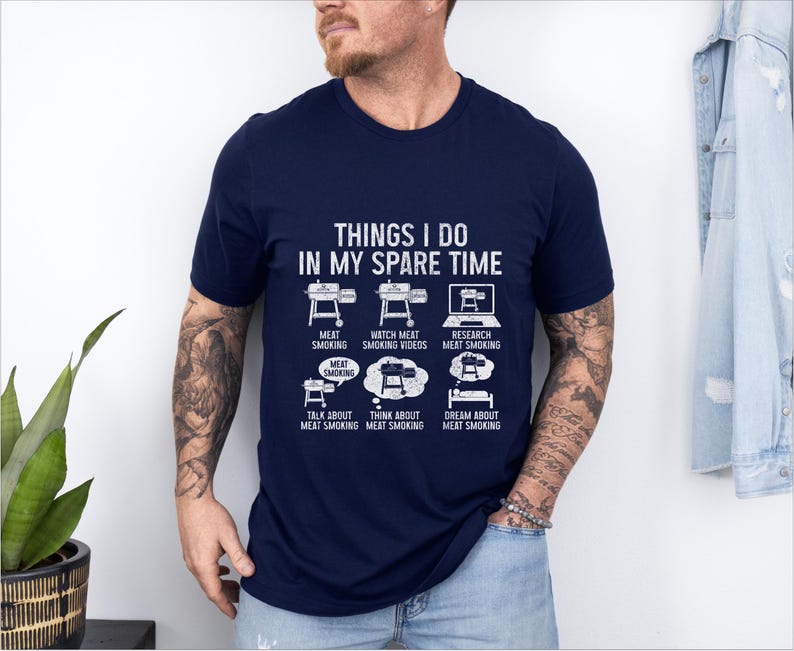 May include: Navy blue t-shirt with white text and graphics. The shirt reads "THINGS I DO IN MY SPARE TIME" with illustrations of a smoker, laptop, and thought bubbles. The graphics depict activities related to meat smoking.