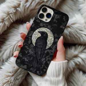 May include: A black smartphone case featuring a detailed illustration of a raven perched on a branch. The bird is centered against a circular, cream-colored stained glass pattern, set against a dark, textured background of foliage and leaves.