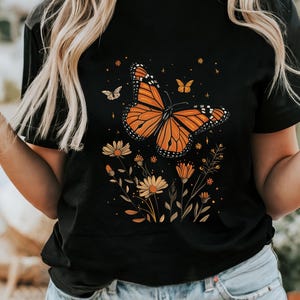 K&ouml;nnte beinhalten: Schwarzes T-Shirt mit einem Aufdruck eines orangefarbenen Schmetterlings und gelber Blumen.