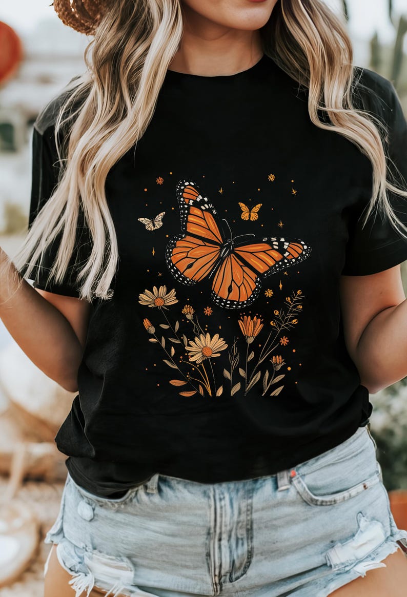 K&ouml;nnte beinhalten: Schwarzes T-Shirt mit einem Aufdruck eines orangefarbenen Schmetterlings und gelber Blumen.
