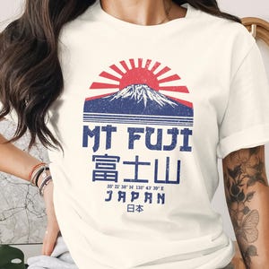 Può includere: T-shirt color crema con grafica del Monte Fuji con un sole rosso, montagna blu e testo. Il testo include "MT FUJI" in inglese e caratteri giapponesi, più coordinate e la parola "JAPAN".
