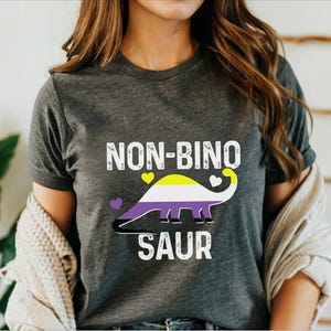 May include: Dark gray t-shirt with the text "NON-BINO SAUR" in white. The design features a dinosaur with the non-binary pride flag colors: yellow, white, purple, and black. Two yellow hearts and one white heart are also included.
