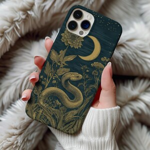Puede incluir: Una funda de teléfono con un fondo verde azulado oscuro que presenta una ilustración dorada de una serpiente, una luna y elementos florales. La funda tiene un acabado mate y un diseño detallado, con una estética vintage.