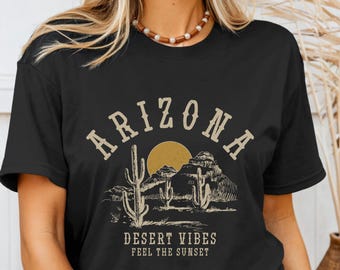 May include: Black t-shirt with a graphic design of a desert landscape with the text "Arizona Desert Vibes Feel the Sunset".