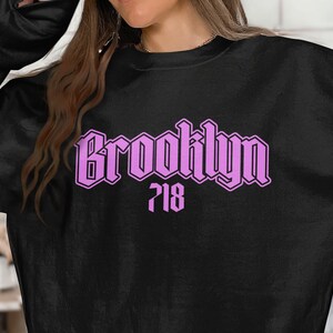 May include: Black sweatshirt with the word "Brooklyn" in a light purple gothic font, with the number "718" below it. The sweatshirt is a crew neck style.
