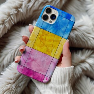 May include: A mobile phone case with a vibrant, geometric design. The case is divided into blue, yellow, and pink sections. The case is held in a hand, with a soft, fluffy background.