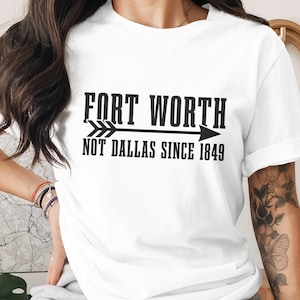 May include: A white short-sleeved t-shirt featuring black text that reads FORT WORTH NOT DALLAS SINCE 1849, with a horizontal arrow graphic positioned between the two lines of text. The shirt has a crew neckline and a standard fit.