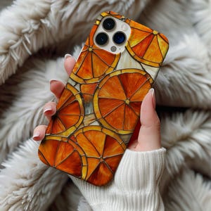 Puede incluir: Una funda de teléfono con un diseño vibrante de rodajas de naranja. La funda tiene un acabado brillante y un patrón repetido de cítricos naranjas y amarillos. La funda se sostiene en la mano de una persona, con un fondo suave y texturizado.