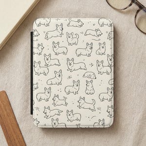 May include: Cream-colored tablet case featuring a repeating pattern of black line-drawn corgi dogs in various poses. The case has a black border and is shown open, with a book and glasses in the background.
