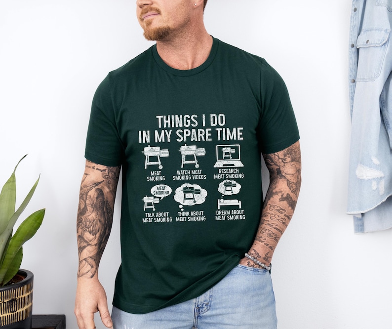 May include: Dark green t-shirt with the text "THINGS I DO IN MY SPARE TIME" and illustrations of meat smoking activities. The shirt features images of smokers, a laptop, and thought bubbles.