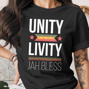Può includere: T-shirt nera con le parole bianche "UNITY AND LIVITY". Il design include una striscia orizzontale colorata e le parole bianche "JAH BLESS". La maglietta ha maniche corte.