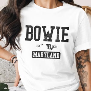 May include: White t-shirt with the word "BOWIE" in large, distressed black letters. Below, it reads "EST. 1870" with a Maryland outline, and "MARYLAND" in a rectangular box. The shirt has rolled-up sleeves.