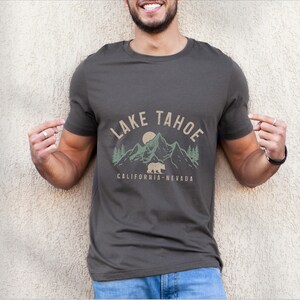 Puede incluir: Una camiseta gris oscuro con un dise&ntilde;o gr&aacute;fico que presenta una cordillera, un oso y el texto "LAKE TAHOE CALIFORNIA-NEVADA".