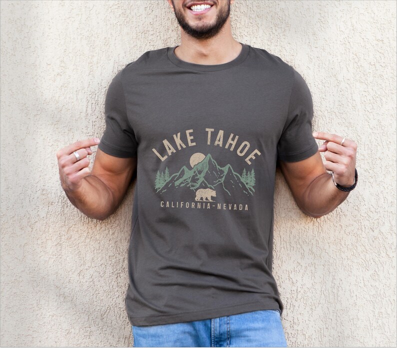Puede incluir: Una camiseta gris oscuro con un dise&ntilde;o gr&aacute;fico que presenta una cordillera, un oso y el texto "LAKE TAHOE CALIFORNIA-NEVADA".