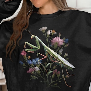 Pode incluir: Camisola preta com uma ilustração detalhada de um louva-a-deus com asas verdes e brancas, num fundo de flores coloridas. As flores incluem flores rosa, azuis e brancas. O desenho está centrado na frente da camisola.