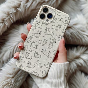 May include: A cream-colored phone case featuring a repeating pattern of black line-drawn corgi dogs in various poses. The case has a camera cutout and is held against a soft, textured background.