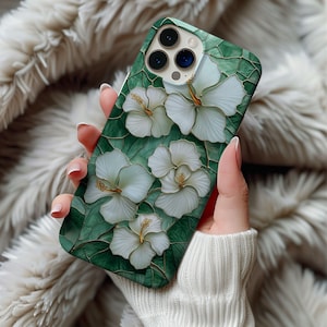 May include: A green phone case featuring white hibiscus flowers with gold accents. The case has a mosaic-like design with a glossy finish. The phone case is held in a person's hand.