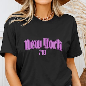 May include: Black t-shirt with the text "New York" in a purple, stylized font, and the number "718" below it. The shirt is a classic crew neck style.