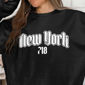 May include: Black sweatshirt with the words "New York" in a white, gothic-style font, with the number "718" below. The sweatshirt is a solid black color.