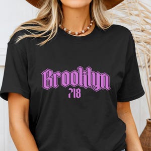 May include: Black t-shirt with the word "Brooklyn" in a purple, stylized font, and the number "718" below it. The shirt is a crew neck style.