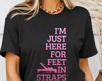 May include: Black t-shirt with the text "I'M JUST HERE FOR FEET IN STRAPS" in pink. The graphic includes a pink illustration of feet in straps. The shirt has short sleeves and a crew neck.