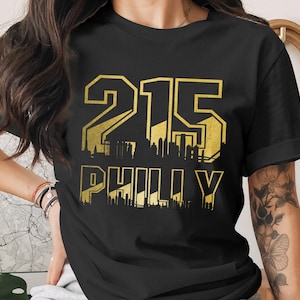May include: Black t-shirt with a gold graphic design. The design features the number "215" above the word "PHILLY" in a bold, stylized font. The design includes a cityscape silhouette.