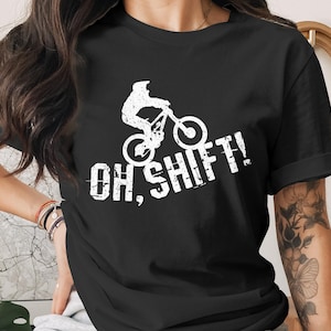 Könnte beinhalten: Schwarzes T-Shirt mit einer weißen Grafik eines Mountainbikers und dem Text "OH, SHIFT!". Das Design hat einen Used-Look mit Textur. Das Shirt ist ein klassischer Rundhalsausschnitt.