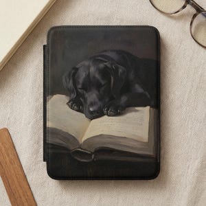 May include: Black e-reader cover featuring a painting of a black Labrador Retriever sleeping on an open book. The cover is rectangular and has a dark, textured appearance. The book's pages are aged, with visible text.