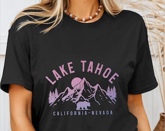 May include: Black t-shirt featuring a graphic design with the words "Lake Tahoe" in pink and purple, along with a mountain range, sun, bear, and trees. The text "California-Nevada" is below the graphic.