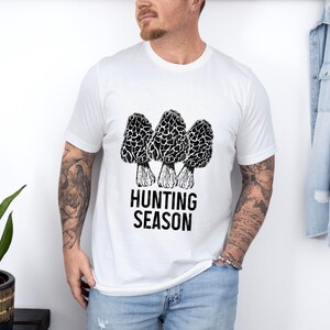 May include: White t-shirt with a black and white illustration of three morel mushrooms and the text "Hunting Season".