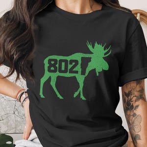 May include: Black t-shirt featuring a green moose silhouette with the number "802" inside. The moose design has a distressed, vintage look. The shirt is a casual wear item.