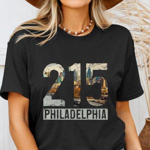 May include: Black t-shirt featuring the number "215" with a cityscape design, and the word "PHILADELPHIA" in a white rectangular box. The shirt is a classic crew neck style.