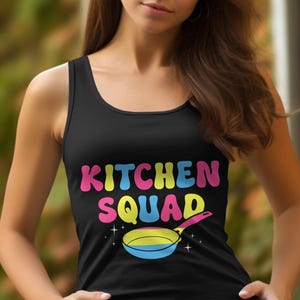 May include: Black vest top featuring the text "KITCHEN SQUAD" in vibrant, bubble-style lettering. A cartoon frying pan with a pink handle and a blue, yellow, and pink pan is below the text. The top is worn by a person.