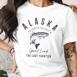 Puede incluir: Camiseta blanca con un diseño gráfico que presenta la palabra "ALASKA" sobre una ilustración de un pez. Las coordenadas 64.2008°N 149.4937°W están impresas sobre el pez. Debajo, las palabras "Great Land" y "THE LAST FRONTIER".