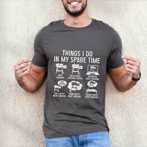 May include: Dark gray t-shirt with white text and graphics. The shirt reads "THINGS I DO IN MY SPARE TIME" with illustrations of a smoker, laptop, and thought bubbles related to meat smoking. A fun, casual tee for barbecue enthusiasts.