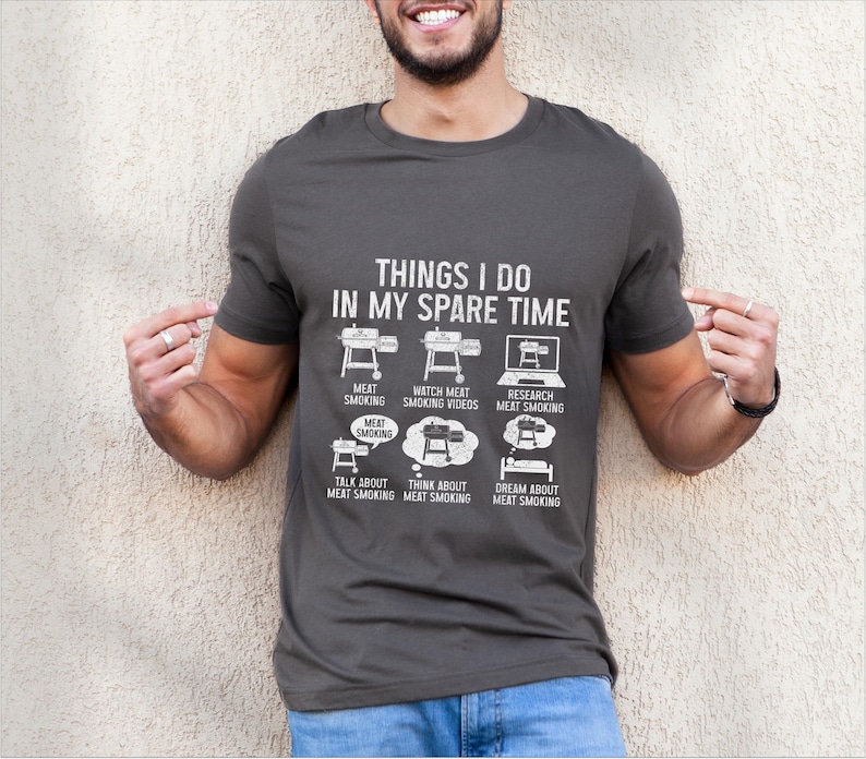 May include: Dark gray t-shirt with white text and graphics. The shirt reads "THINGS I DO IN MY SPARE TIME" with illustrations of a smoker, laptop, and thought bubbles related to meat smoking. A fun, casual tee for barbecue enthusiasts.