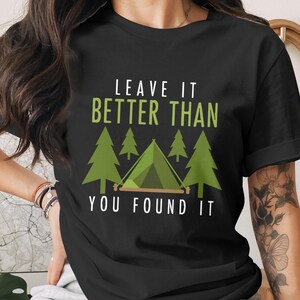 May include: Black t-shirt with the text "LEAVE IT BETTER THAN YOU FOUND IT" in white and green. The design includes a green tent and pine trees, perfect for camping enthusiasts.