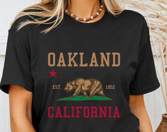 May include: Black t-shirt featuring the text "OAKLAND" in a tan font above a brown bear graphic. Below the bear is the word "CALIFORNIA" in red. A red star and the text "EST. 1852" are also present.