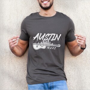 May include: A dark gray t-shirt with a white graphic design of the Austin, Texas skyline with a guitar. The text "Austin Texas" is printed below the skyline.