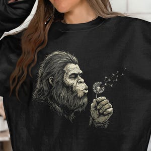 May include: A black long-sleeve sweatshirt featuring a graphic illustration of a hairy, ape-like creature blowing on a dandelion. The creature is depicted in shades of grey and white, with the dandelion seeds drifting away into the air.