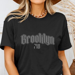 May include: Black t-shirt with the word "Brooklyn" in a gray, gothic-style font, and the number "718" below it. The shirt is a crew neck and appears to be made of cotton. The model is wearing a brown hat and a beaded necklace.