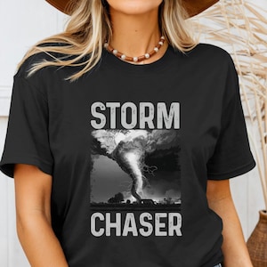 May include: Black t-shirt with the words "STORM CHASER" in white block letters above and below a grayscale image of a tornado. The image shows a swirling funnel cloud over a rural landscape.