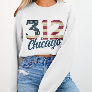May include: White sweatshirt with the number "312" in a distressed American flag design, and the word "Chicago" in a navy blue script font. The sweatshirt is paired with blue jeans.