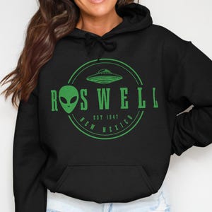 May include: A black hooded sweatshirt featuring a green graphic print on the chest. The design includes a circular border, a flying saucer, an alien head, and the text ROSWELL NEW MEXICO EST 1947. The hoodie has long sleeves and a front pouch pocket.