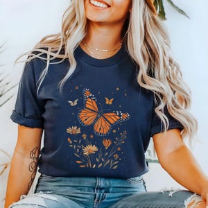 K&ouml;nnte beinhalten: Ein marineblaues T-Shirt mit einem Aufdruck eines orange-schwarzen Schmetterlings, umgeben von gelben Blumen und gr&uuml;nen Bl&auml;ttern. Der Schmetterling hat ein detailliertes Design und ist vor einem dunkelblauen Hintergrund platziert.